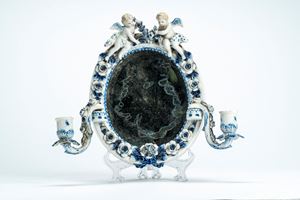 Specchierina di forma ovale in porcellana decorata a fiori, foglie, fiocco a rilievo e sormontata da due putti alati con festone, con due bracci porta candela removibili a voluta. Manifattura tedesca, secolo XIX