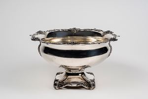 Grande coppa centrotavola in metallo argentato su quattro piedi a volute fogliate con bordo mosso a sei mascheroni e girali di foglie